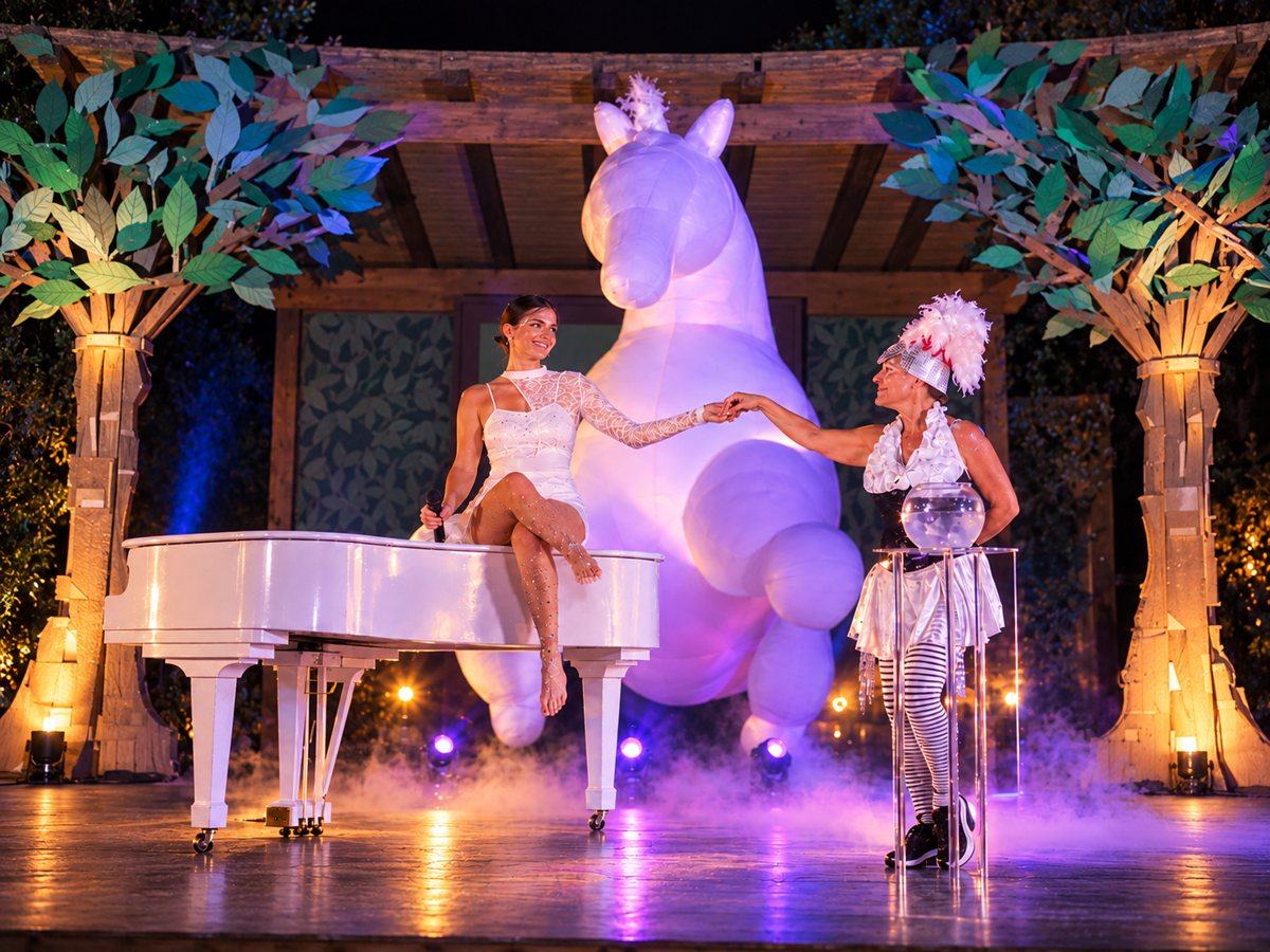 Cantante seduta su un pianoforte bianco e artista in costume con pennacchio, davanti a un grande unicorno luminoso